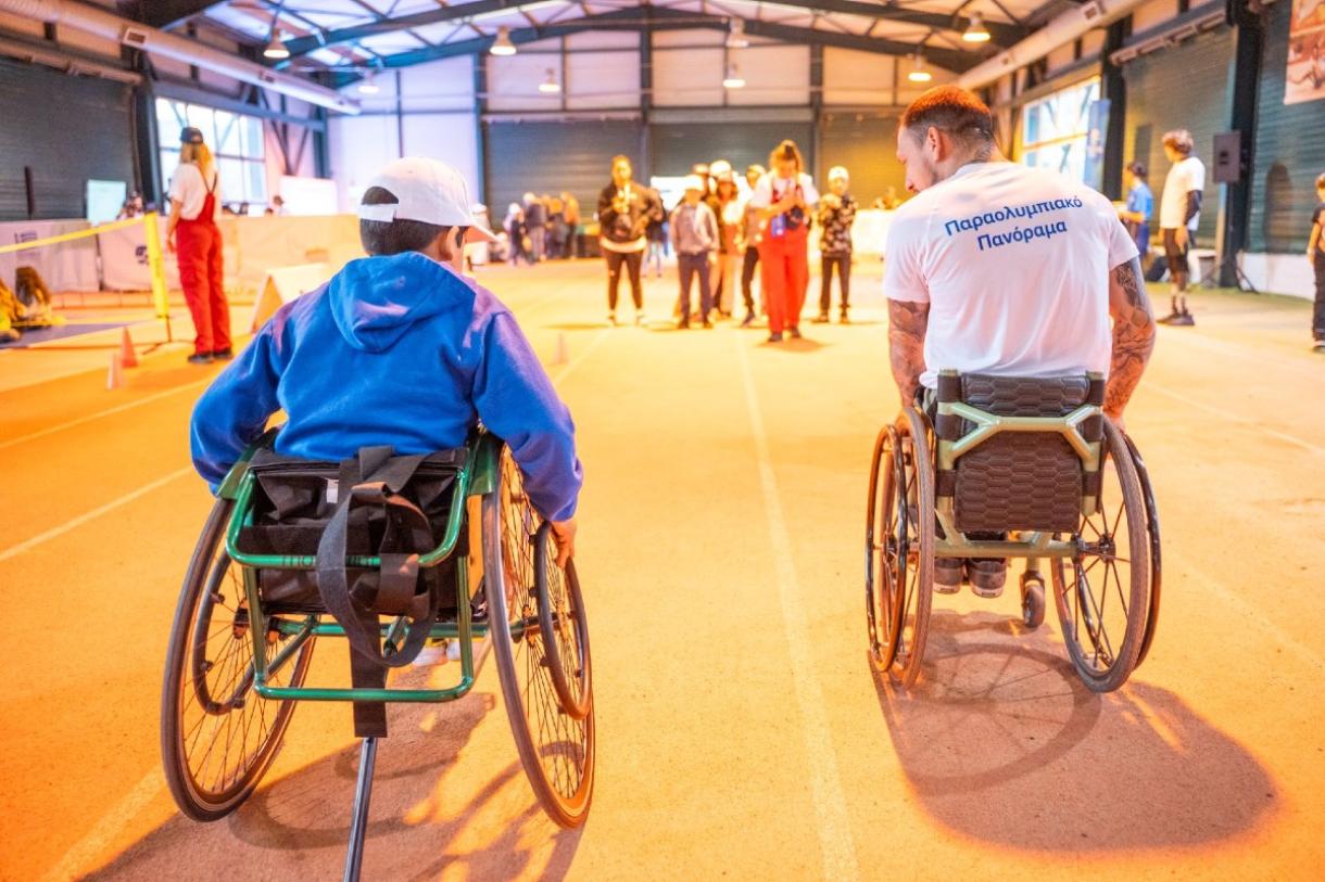 ένα παιδί και ένας ενήλικας σε αγωνιστικά αναπηρικά αμαξίδια, κινούνται σε κλειστό στίβο προς μια ομάδα ανθρώπων